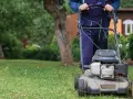 Mężczyzna w niebieskich ogrodniczkach kosi trawnik. Widać skoszoną, suchą trawę po pracy kosiarki.